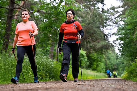 Zwei Frauen gehen mit Nordic-Walking-Stöcken auf einem Waldweg, umgeben von Bäumen und grüner Vegetation.